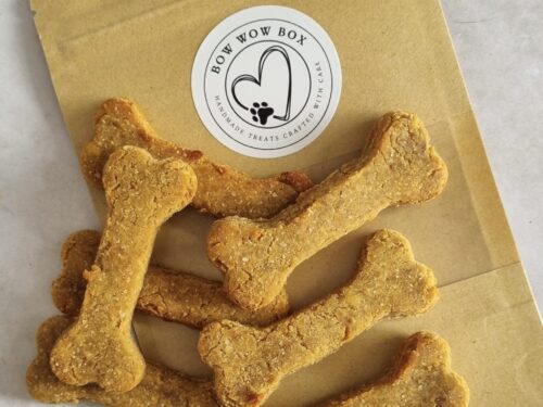 Bone-shaped dog treats spilling from a brown 'Bow Wow Box' branded bag with a paw print logo, highlighting handmade, crafted treats.
