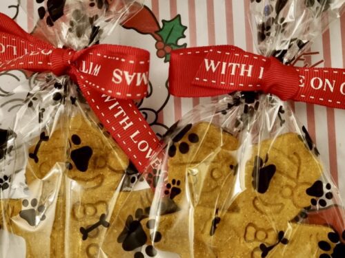 Gingerbread-person shaped dog treats wrapped in cellophane bags with red festive ribbon saying "with love" and paw print wrapping paper background