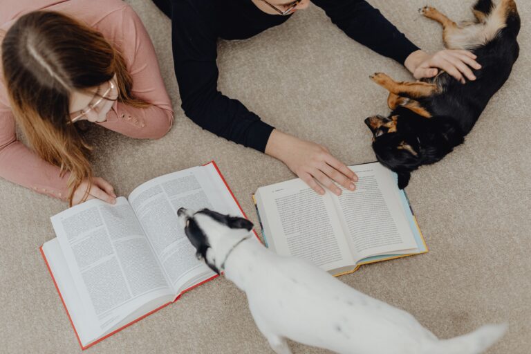 Two people lying on the floor reading books while two dogs rest beside them, illustrating a calm back-to-school routine for dogs