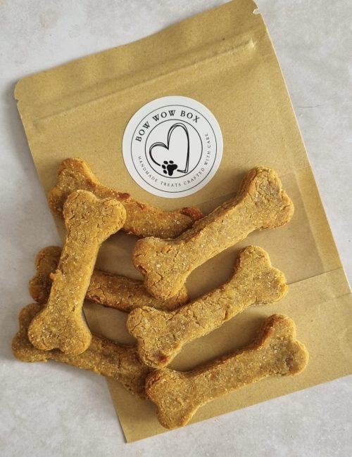 Bone-shaped dog treats spilling from a brown 'Bow Wow Box' branded bag with a paw print logo, highlighting handmade, crafted treats.